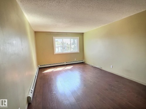 Spacious room featuring wood-finish flooring, a large window with white trim, and baseboard heating - 306 9730 156 Street, Edmonton, AB 