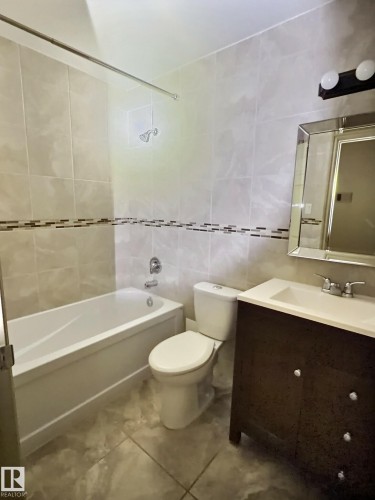 Bathroom featuring light-toned wall tiling with a decorative accent band, a white soaking tub with a shower fixture, a white toilet, and a dark wood-finish vanity with an integrated sink - 306 9730 156 Street, Edmonton, AB 