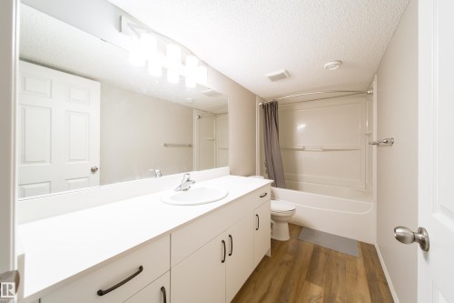 Bathroom featuring a full vanity with a white countertop, an integrated sink, a large wall-mounted mirror, a tub and shower combination with white surround, and wood-finish flooring - 3038 Checknita Way Sw, Edmonton, AB 