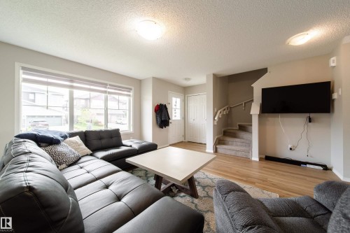 Bright interior space featuring a large window with horizontal blinds, wood-finish flooring, a carpeted staircase with a white railing, a white entry door, and flush-mount ceiling lighting - 3038 Checknita Way Sw, Edmonton, AB 
