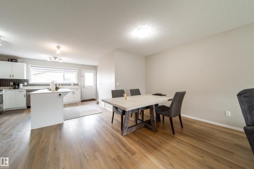 Open-concept living space featuring wood-finish flooring, a kitchen island, white cabinetry, dark tile backsplash, and stainless steel appliances - 3038 Checknita Way Sw, Edmonton, AB 