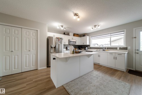 Bright kitchen featuring a central island with a light-toned countertop, white cabinetry, a dark tile backsplash, stainless steel appliances, and wood-finish flooring - 3038 Checknita Way Sw, Edmonton, AB 