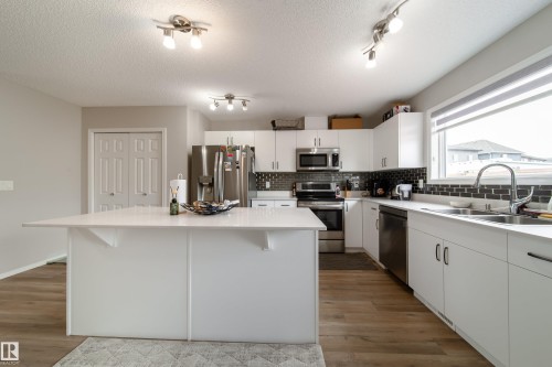 Bright kitchen featuring a central island with a solid surface countertop, stainless steel appliances, white cabinetry, wood-finish flooring, and a tile backsplash - 3038 Checknita Way Sw, Edmonton, AB 