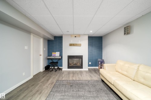Living area featuring a built-in fireplace with a wood mantel, wood-finish flooring, a drop ceiling, and a distinct accent wall - 8512 142A Avenue, Edmonton, AB 