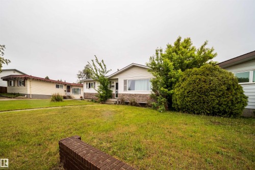 Expansive front lawn with mature landscaping, a brick garden wall, and a single-story residence featuring white siding and a stone-clad foundation - 8512 142A Avenue, Edmonton, AB 