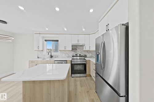 Modern kitchen featuring a large island with a light-toned countertop, stainless steel appliances, white upper cabinetry, and wood-finish lower cabinetry - 6144 166 Avenue, Edmonton, AB 