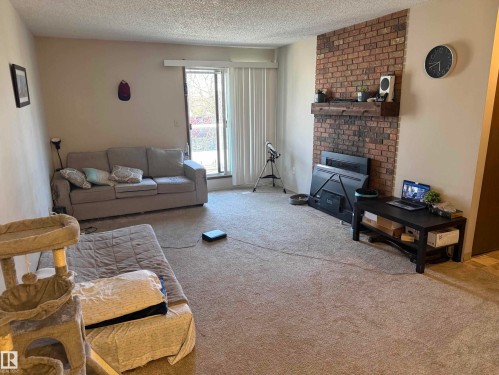 Living area featuring a brick accent wall with a wooden mantel, light-colored carpeting, and a sliding glass door providing access to the outdoors - 304 10604 110 Avenue, Edmonton, AB 