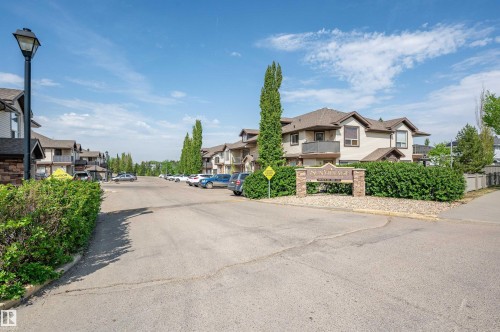 Paved roadway with a stone and wood-finish entry sign, surrounded by mature hedges and residential buildings featuring balconies and multi-pane windows - 40 604 62 Street, Edmonton, AB 