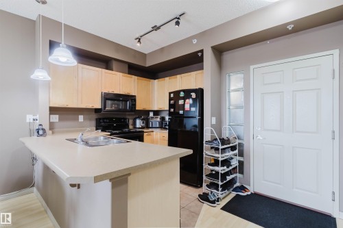 Kitchen featuring light wood cabinetry, black appliances, track lighting, and a peninsula with an integrated sink - 1-405 4245 139 Avenue, Edmonton, AB 
