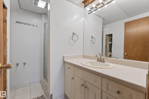Bathroom featuring an integrated vanity sink with a brushed nickel faucet, a spacious mirror with overhead bar lighting, a shower enclosure with a glass door, and a tiled floor - 6203 90 Avenue, Edmonton, AB 
