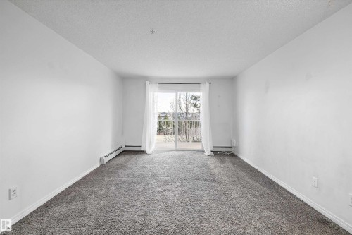 Spacious room featuring gray textured carpet, white painted walls, and a sliding glass door leading to an outdoor area with a white railing - 320 245 Edwards Drive, Edmonton, AB 