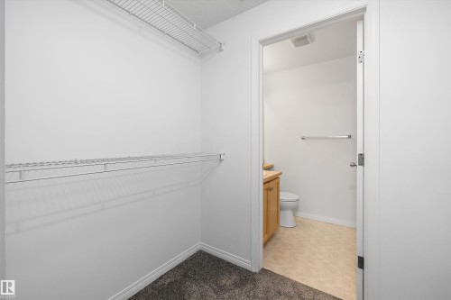 Walk-in closet featuring wire shelving and dark gray carpeting, with a partial bathroom entrance showcasing a wood-finish vanity and beige flooring - 320 245 Edwards Drive, Edmonton, AB 