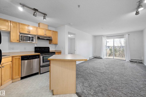 Open-concept living area with a kitchen featuring natural wood cabinetry, stainless steel appliances, and a central island - 320 245 Edwards Drive, Edmonton, AB 