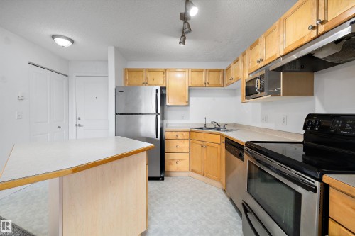 Kitchen featuring wood cabinetry, stainless steel appliances, a corner sink, a built-in microwave, and track lighting - 320 245 Edwards Drive, Edmonton, AB 