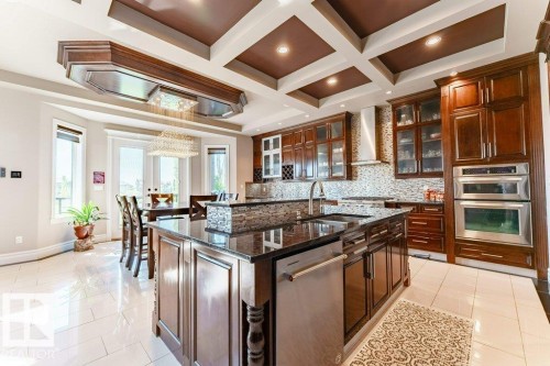 Gourmet kitchen featuring a large central island with a dark countertop, rich wood cabinetry, stainless steel appliances, a mosaic tile backsplash, and a coffered ceiling - 328 52327 Range Rd 233, Rural Strathcona County, AB 