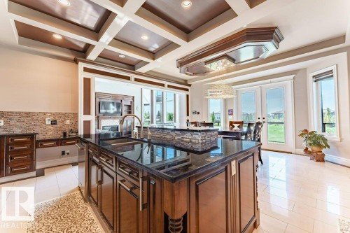 Kitchen featuring a large central island with a dark countertop and integrated sink, coffered ceiling, light-toned flooring, and a stone-tiled backsplash - 328 52327 Range Rd 233, Rural Strathcona County, AB 