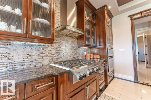 Gourmet kitchen featuring a six-burner gas range with stainless steel hood, built-in double ovens, dark wood cabinetry with glass-front uppers, and a mosaic tile backsplash - 328 52327 Range Rd 233, Rural Strathcona County, AB 
