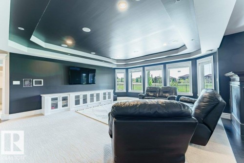 Spacious entertainment room featuring a coffered ceiling with recessed lighting, an expansive built-in white media console with glass-front cabinetry, a wall-mounted television, and a bay window wall with multiple windows - 328 52327 Range Rd 233, Rural Strathcona County, AB 