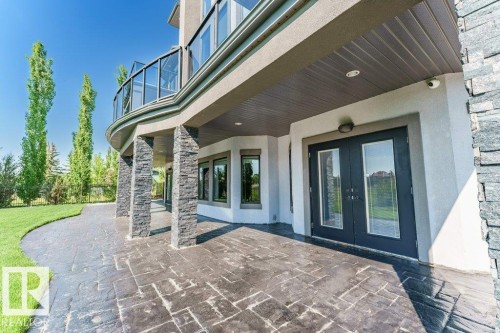 Expansive stamped concrete patio featuring stone-clad support pillars, a manicured lawn, and direct access through double glass-paneled doors - 328 52327 Range Rd 233, Rural Strathcona County, AB 