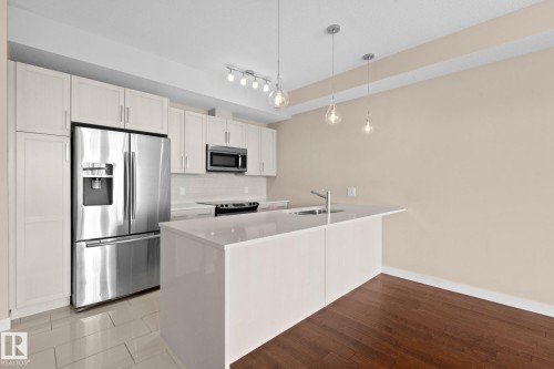 Modern kitchen featuring a waterfall island, stainless steel appliances, white cabinetry, and wood-finish flooring - 309 625 Leger Way, Edmonton, AB 