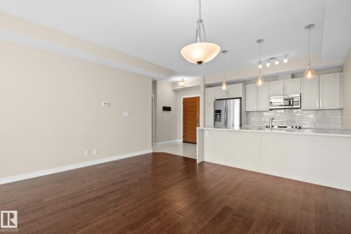 Open-concept living space featuring wood-finish flooring, a modern kitchen island, stainless steel appliances, a subway tile backsplash, and contemporary pendant lighting - 309 625 Leger Way, Edmonton, AB 