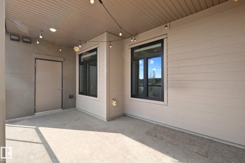Covered outdoor patio featuring a concrete floor, two windows with dark frames, and string lighting - 309 625 Leger Way, Edmonton, AB 