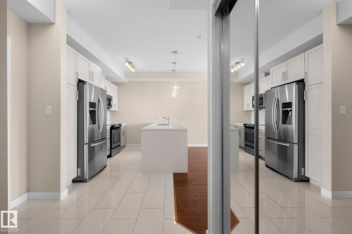 Modern kitchen featuring an expansive center island, stainless steel appliances, white cabinetry, track lighting, and light-toned tile flooring - 309 625 Leger Way, Edmonton, AB 