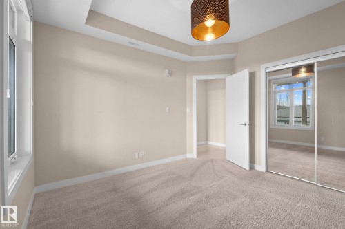 Carpeted room featuring a tray ceiling, a drum pendant light fixture, a window with white trim, and mirrored sliding closet doors - 309 625 Leger Way, Edmonton, AB 