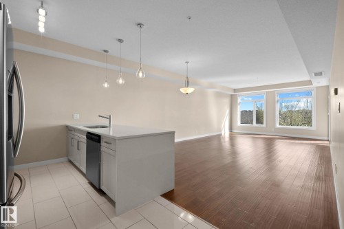 Open-concept living area featuring wood-finish flooring, a kitchen island with a built-in sink and dishwasher, pendant lighting, and multiple windows - 309 625 Leger Way, Edmonton, AB 