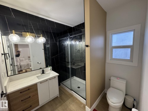 Modern bathroom featuring a vanity with a white countertop and wood-finish drawers, black-veined wall tiling, a walk-in shower with a glass enclosure, and wood-finish flooring - 2123 18 Avenue, Edmonton, AB 