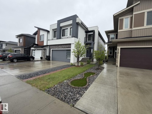 Modern exterior featuring white and dark gray stucco, stone veneer accents, an attached garage with a gray door, a second-story balcony with dark railings, and a landscaped front yard with grass and decorative rock - 2123 18 Avenue, Edmonton, AB 