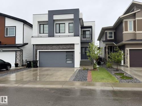 Modern multi-level facade featuring contrasting white and gray siding, stone veneer accents, a dark gray garage door with horizontal windows, and a second-story balcony with dark railings - 2123 18 Avenue, Edmonton, AB 