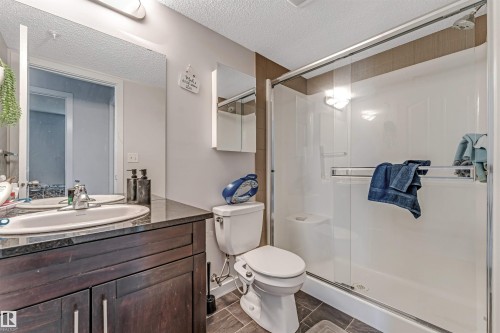 Bathroom featuring a dark wood-finish vanity with an undermount sink and dark countertop, a large wall-mounted mirror, and a glass-enclosed shower with white paneling and a built-in shelf - 320 534 Watt Boulevard, Edmonton, AB - Indoor Photo Showing Bathroom