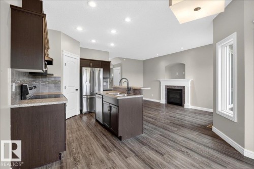 Open-concept living area featuring wood-finish flooring, a kitchen island with sink and integrated dishwasher, dark wood cabinetry, stainless steel refrigerator, and a fireplace with a white mantel and tile surround - 62 Durrand Bend, Fort Saskatchewan, AB 