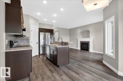 Open-concept living area featuring wood-finish flooring, a kitchen island with sink and integrated dishwasher, dark wood cabinetry, stainless steel refrigerator, and a fireplace with a white mantel and tile surround - 