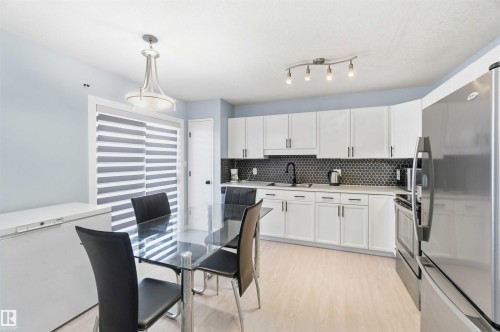Modern kitchen featuring white cabinetry, a dark hexagonal tile backsplash, light wood-finish flooring, a stainless steel refrigerator, and a sliding glass door - 8 15128 22 Street, Edmonton, AB 