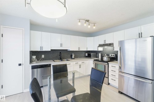 Modern kitchen featuring white cabinetry, a dark hexagonal tile backsplash, stainless steel appliances, light wood-finish flooring, and a contemporary ceiling light fixture - 8 15128 22 Street, Edmonton, AB 