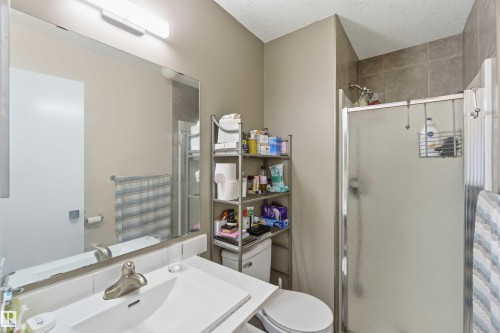 Bathroom featuring a vanity with an integrated sink, a large wall-mounted mirror, overhead bar lighting, a commode, and a shower enclosure with a tiled surround - 8 15128 22 Street, Edmonton, AB 