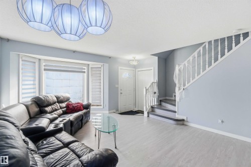 Bright living area featuring light wood-finish flooring, a bay window with horizontal blinds, and a contemporary ceiling light fixture - 8 15128 22 Street, Edmonton, AB 