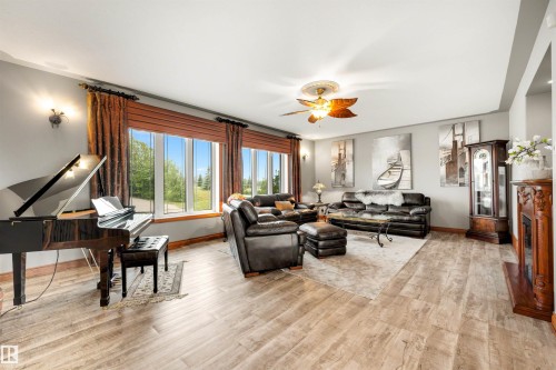 Spacious living area featuring large windows, light wood-finish flooring, a decorative ceiling fan, and a built-in fireplace with an ornate wooden mantel - 28125 Highway 587, Rural Red Deer County, AB 