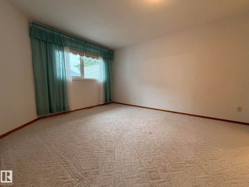 Room featuring light-toned patterned carpeting, neutral wall finishes, and a single window with horizontal blinds and teal-colored drapes - 102 4610 50 Avenue, Stony Plain, AB 