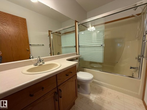 Bathroom featuring a single vanity with an integrated sink, a full-width mirror, and a tub-shower combination with a sliding glass enclosure - 102 4610 50 Avenue, Stony Plain, AB 