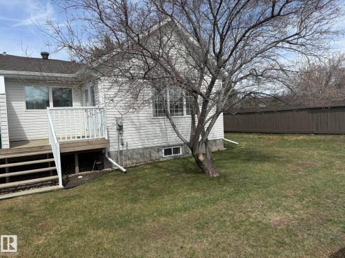 Rear exterior featuring white vinyl siding, a wood deck with white railings, a mature deciduous tree, and a brown privacy fence - 102 4610 50 Avenue, Stony Plain, AB 
