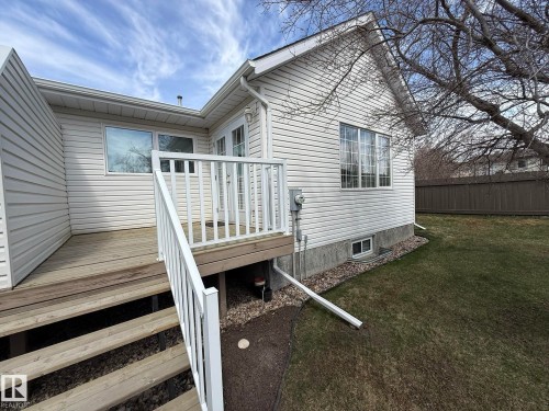 Rear exterior featuring a wooden deck with white railing, vinyl siding, multiple windows, and a grassy yard with a privacy fence - 102 4610 50 Avenue, Stony Plain, AB 