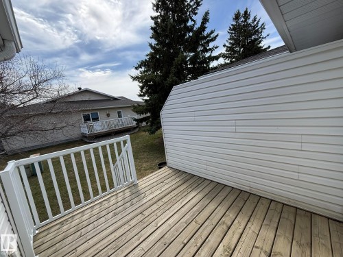 Wood-finish deck with white railing, featuring white siding and evergreen trees in the background - 102 4610 50 Avenue, Stony Plain, AB 