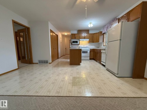 Kitchen featuring wood cabinetry, white appliances, a central island, and patterned vinyl flooring - 102 4610 50 Avenue, Stony Plain, AB 