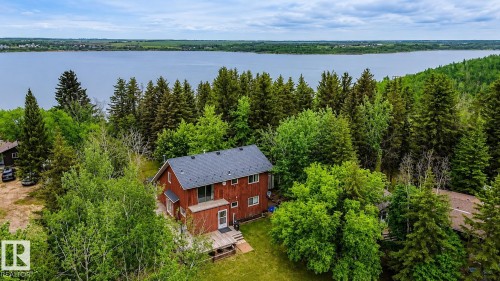 Red-sided residence featuring a dark gray roofline, surrounded by dense evergreen and deciduous trees, with a prominent body of water in the background - 61 Stang Cres, Rural Two Hills County, AB 
