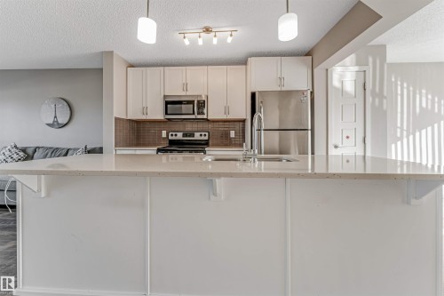 Kitchen island featuring a light-toned countertop, white cabinetry, stainless steel appliances, a tile backsplash, and overhead pendant lighting - 1624 33A Street, Edmonton, AB 