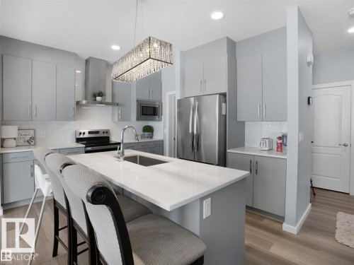 Modern kitchen featuring a large center island with seating, stainless steel appliances, light gray cabinetry, white subway tile backsplash, and wood-finish flooring - 207 41 Avenue, Edmonton, AB 