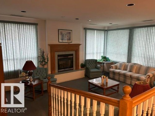 Expansive living area featuring a central fireplace with an ornate wood mantel, recessed ceiling lighting, a large window array with vertical blinds, and a wood railing with white balusters - 50 303 Twin Brooks Drive Nw, Edmonton, AB 
